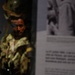 A museum display featuring a military mannequin dressed in camouflage combat gear, positioned behind glass in a dimly lit exhibit case. To the right, an informational panel with text and a black-and-white archival photograph is visible, though both are partially out of focus. The exhibit appears to document military history, likely related to a specific conflict or soldier's experience.
