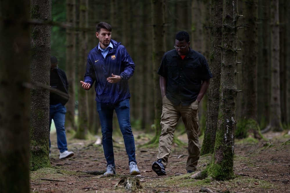 Three individuals are walking through a dense, evenly-spaced forest with tall, slender trees and moss-covered ground. Two men are prominently visible in the foreground — one wearing a blue FC Barcelona jacket and jeans, appearing to be speaking, and another in a dark shirt and khaki cargo pants looking downward. A third person is partially visible in the background on the left.