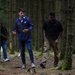 Three individuals are walking through a dense, evenly-spaced forest with tall, slender trees and moss-covered ground. Two men are prominently visible in the foreground — one wearing a blue FC Barcelona jacket and jeans, appearing to be speaking, and another in a dark shirt and khaki cargo pants looking downward. A third person is partially visible in the background on the left.