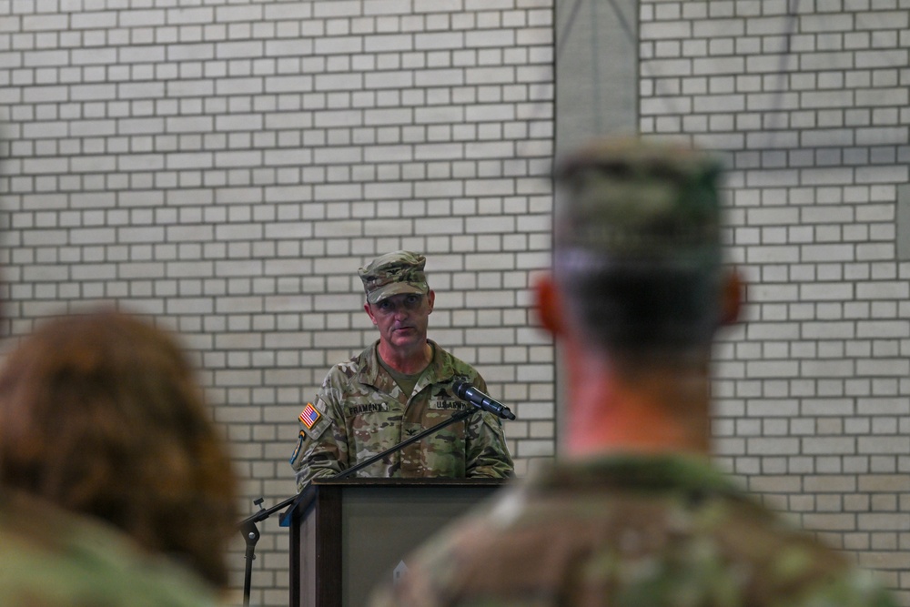 A U.S. Army soldier in Operational Camouflage Pattern (OCP) uniform is speaking at a podium with a microphone during what appears to be a formal military ceremony or briefing. The speaker's name tape reads 'FRAMONT' (or similar) and a U.S. flag patch is visible on the right shoulder. An audience of uniformed military personnel is visible in the foreground, out of focus.