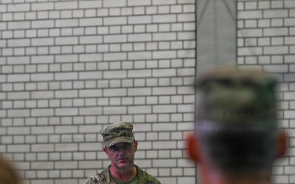 A U.S. Army soldier in Operational Camouflage Pattern (OCP) uniform is speaking at a podium with a microphone during what appears to be a formal military ceremony or briefing. The speaker's name tape reads 'FRAMONT' (or similar) and a U.S. flag patch is visible on the right shoulder. An audience of uniformed military personnel is visible in the foreground, out of focus.