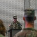 A U.S. Army soldier in Operational Camouflage Pattern (OCP) uniform is speaking at a podium with a microphone during what appears to be a formal military ceremony or briefing. The speaker's name tape reads 'FRAMONT' (or similar) and a U.S. flag patch is visible on the right shoulder. An audience of uniformed military personnel is visible in the foreground, out of focus.