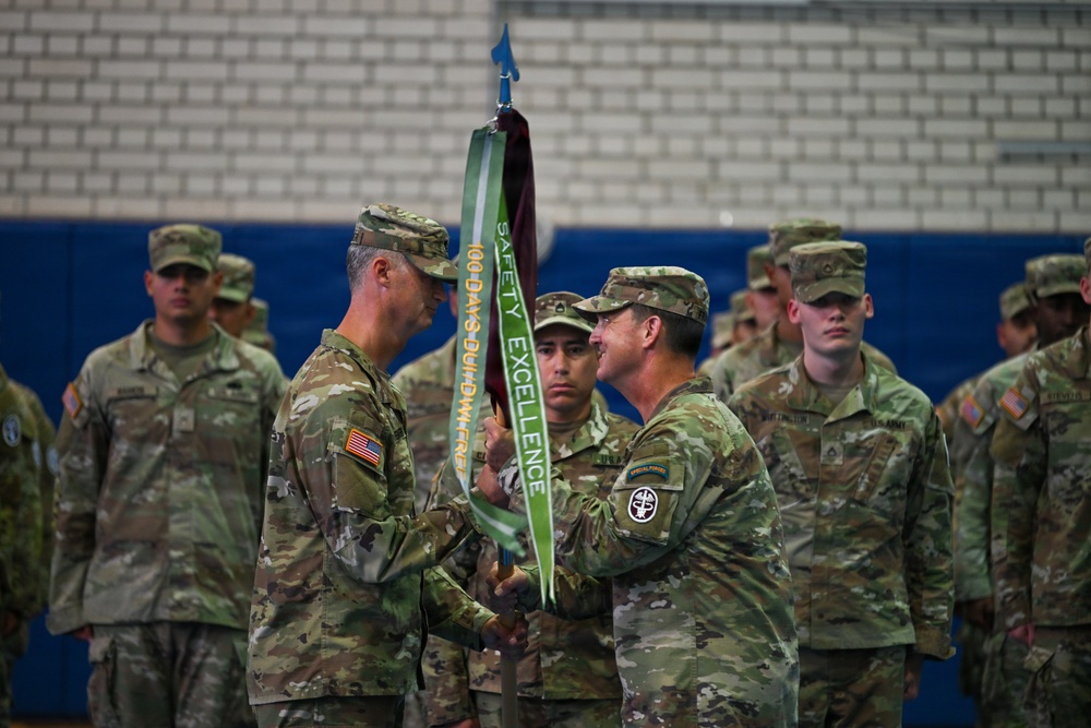 Two senior U.S. Army soldiers are conducting a formal guidon/streamer presentation ceremony indoors, likely in a gymnasium or drill hall. One soldier is passing or receiving a unit guidon staff adorned with a green streamer reading 'SAFETY EXCELLENCE' and '100 DAYS ACCIDENT FREE'. A formation of soldiers stands at attention in the background as witnesses to the ceremony.