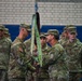 Two senior U.S. Army soldiers are conducting a formal guidon/streamer presentation ceremony indoors, likely in a gymnasium or drill hall. One soldier is passing or receiving a unit guidon staff adorned with a green streamer reading 'SAFETY EXCELLENCE' and '100 DAYS ACCIDENT FREE'. A formation of soldiers stands at attention in the background as witnesses to the ceremony.