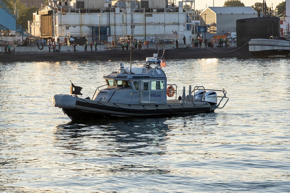 A U.S. Navy Police patrol boat is operating on a waterway near a naval installation. In the background, a submarine is docked at a pier with numerous personnel visible on the dock. The scene appears to be at a naval base, likely during a significant event given the crowd of people present.