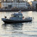A U.S. Navy Police patrol boat is operating on a waterway near a naval installation. In the background, a submarine is docked at a pier with numerous personnel visible on the dock. The scene appears to be at a naval base, likely during a significant event given the crowd of people present.