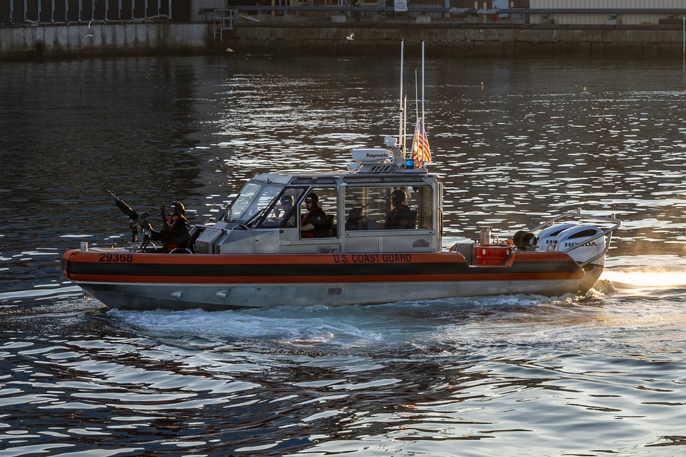 A U.S. Coast Guard patrol boat numbered 29368 is underway on a waterway, likely in a harbor or port area. The vessel is an orange and gray rigid inflatable boat (RIB) with multiple crew members aboard, some appearing to be armed. The image was taken during golden hour lighting conditions with an industrial waterfront structure visible in the background.