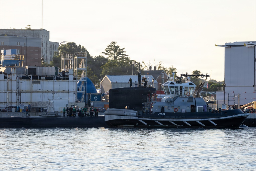 A naval yard scene showing a dark-hulled submarine moored alongside an industrial pier or floating dry dock. A harbor tug boat marked 'YT809' with the name 'MOMENTOUS' is positioned adjacent to the submarine, likely assisting with maneuvering. Several personnel in work gear are visible on the pier and vessels, with industrial shipyard buildings and trees visible in the background.