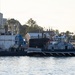 A naval yard scene showing a dark-hulled submarine moored alongside an industrial pier or floating dry dock. A harbor tug boat marked 'YT809' with the name 'MOMENTOUS' is positioned adjacent to the submarine, likely assisting with maneuvering. Several personnel in work gear are visible on the pier and vessels, with industrial shipyard buildings and trees visible in the background.