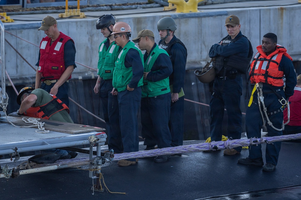 A group of U.S. Navy personnel are gathered on what appears to be a ship's deck during a mooring or line-handling operation. Personnel are wearing a variety of safety gear including helmets, life vests in orange and red, and green safety vests. A large mooring line is visible on the deck, and one individual is crouched down examining equipment near a gangway or brow.
