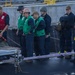 A group of U.S. Navy personnel are gathered on what appears to be a ship's deck during a mooring or line-handling operation. Personnel are wearing a variety of safety gear including helmets, life vests in orange and red, and green safety vests. A large mooring line is visible on the deck, and one individual is crouched down examining equipment near a gangway or brow.