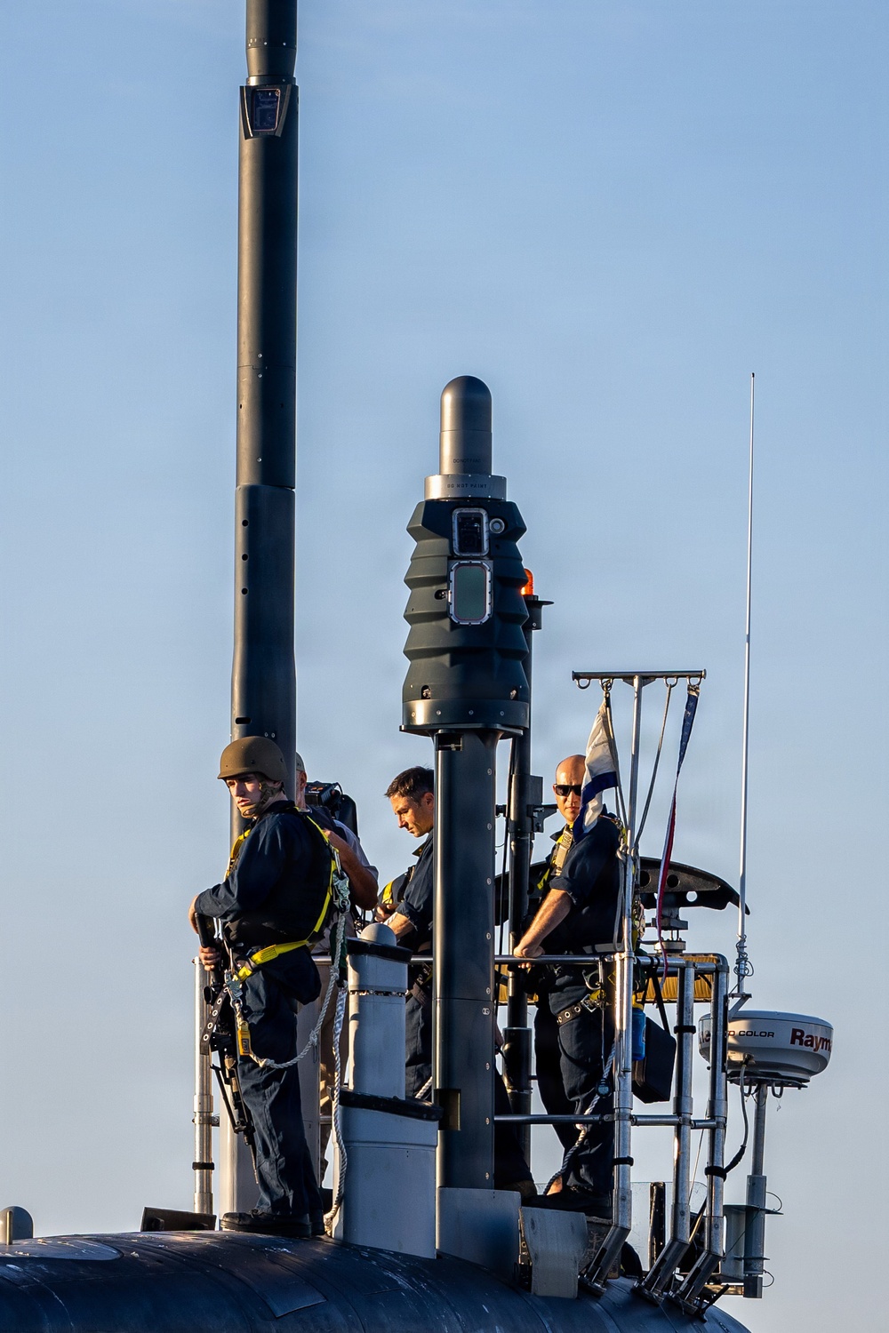 The image shows several naval personnel standing on the sail (conning tower) of a submarine at sea. The crew members are wearing dark uniforms with yellow safety harnesses and are positioned around various mast and periscope structures. The scene appears to be taken during surface transit operations in calm conditions.