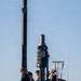 The image shows several naval personnel standing on the sail (conning tower) of a submarine at sea. The crew members are wearing dark uniforms with yellow safety harnesses and are positioned around various mast and periscope structures. The scene appears to be taken during surface transit operations in calm conditions.