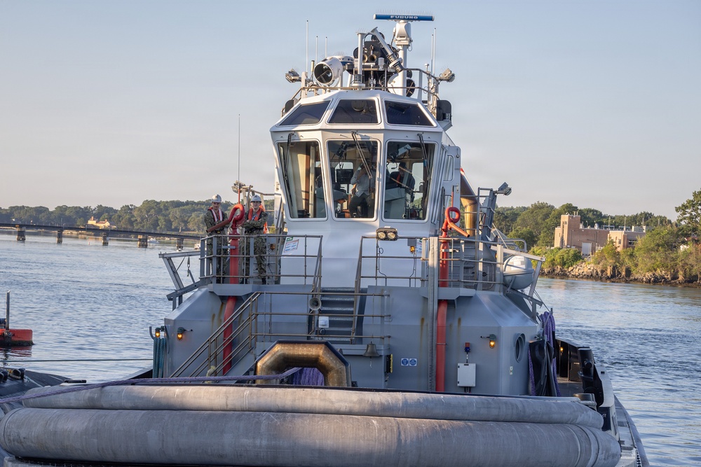 A gray military or government tugboat is operating on a river in what appears to be a harbor or naval facility setting. The vessel features a prominent enclosed wheelhouse, multiple navigation and communication instruments on the mast, and large rubber fendering along the hull. Several crew members in work attire and hard hats are visible on the deck.