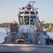 A gray military or government tugboat is operating on a river in what appears to be a harbor or naval facility setting. The vessel features a prominent enclosed wheelhouse, multiple navigation and communication instruments on the mast, and large rubber fendering along the hull. Several crew members in work attire and hard hats are visible on the deck.