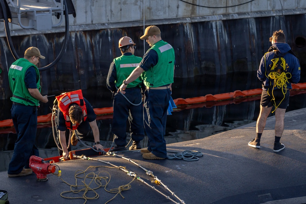 A group of workers on a dock or pier are handling mooring lines and equipment alongside a large vessel. Several personnel wearing green safety vests, a hard hat, and an orange life vest are actively working with ropes and rigging hardware. The scene appears to be a ship berthing or line-handling operation at a maritime facility.