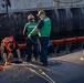 A group of workers on a dock or pier are handling mooring lines and equipment alongside a large vessel. Several personnel wearing green safety vests, a hard hat, and an orange life vest are actively working with ropes and rigging hardware. The scene appears to be a ship berthing or line-handling operation at a maritime facility.