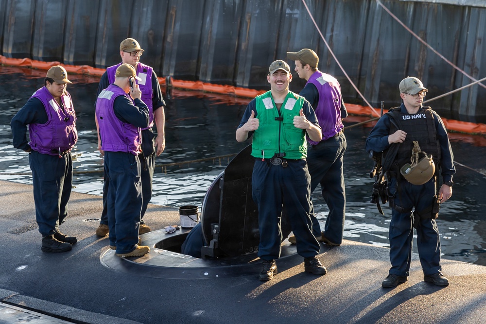A group of U.S. Navy personnel are standing on the deck of what appears to be a submarine, docked near a pier or floating barrier. One sailor in a green vest is giving two thumbs up toward the camera, while others in purple vests and one in a 'US NAVY' jacket stand nearby. The scene appears to be a routine docking or mooring operation.