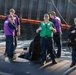 A group of U.S. Navy personnel are standing on the deck of what appears to be a submarine, docked near a pier or floating barrier. One sailor in a green vest is giving two thumbs up toward the camera, while others in purple vests and one in a 'US NAVY' jacket stand nearby. The scene appears to be a routine docking or mooring operation.