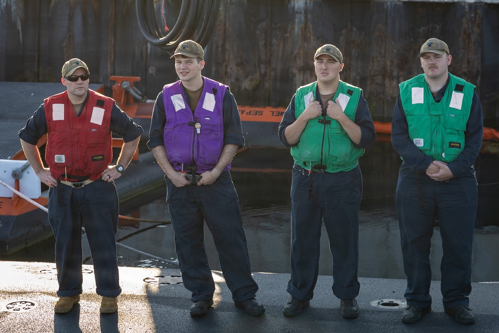 Four personnel stand in a line on what appears to be a naval vessel or dock, each wearing different colored safety/work vests over dark uniforms. They are all wearing matching tan/olive baseball caps and appear to be in a formal or semi-formal standing formation. The setting includes visible maritime equipment and water in the background.