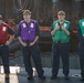 Four personnel stand in a line on what appears to be a naval vessel or dock, each wearing different colored safety/work vests over dark uniforms. They are all wearing matching tan/olive baseball caps and appear to be in a formal or semi-formal standing formation. The setting includes visible maritime equipment and water in the background.