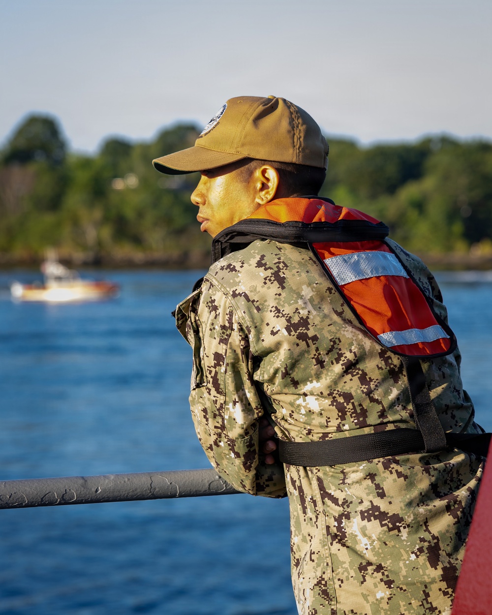A military service member wearing digital camouflage (NWU Type III pattern consistent with U.S. Navy) and an orange life vest stands at a railing, looking out over a body of water. They are wearing a coyote brown cap with a visible patch/emblem. A small boat is visible in the background on the water.
