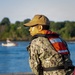 A military service member wearing digital camouflage (NWU Type III pattern consistent with U.S. Navy) and an orange life vest stands at a railing, looking out over a body of water. They are wearing a coyote brown cap with a visible patch/emblem. A small boat is visible in the background on the water.