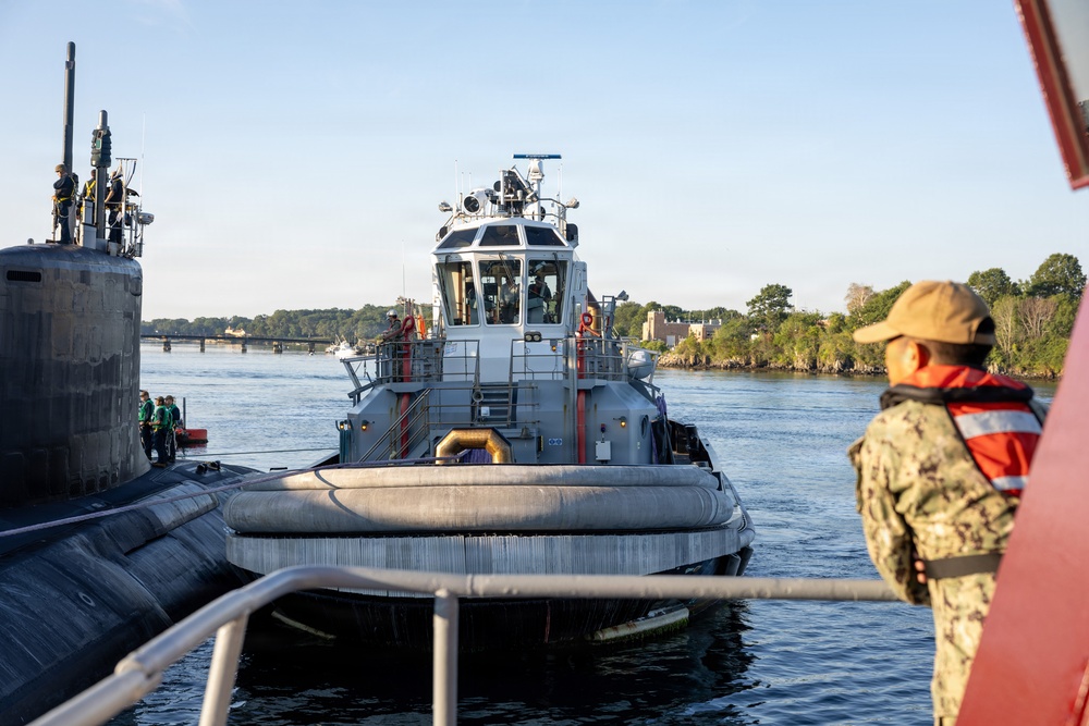 A naval tugboat is positioned alongside a submarine in a harbor or river waterway. A Navy sailor in camouflage uniform and orange life vest observes the operation from the foreground. The scene depicts a standard maritime assist or docking operation at what appears to be a naval installation.