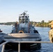 A naval tugboat is positioned alongside a submarine in a harbor or river waterway. A Navy sailor in camouflage uniform and orange life vest observes the operation from the foreground. The scene depicts a standard maritime assist or docking operation at what appears to be a naval installation.