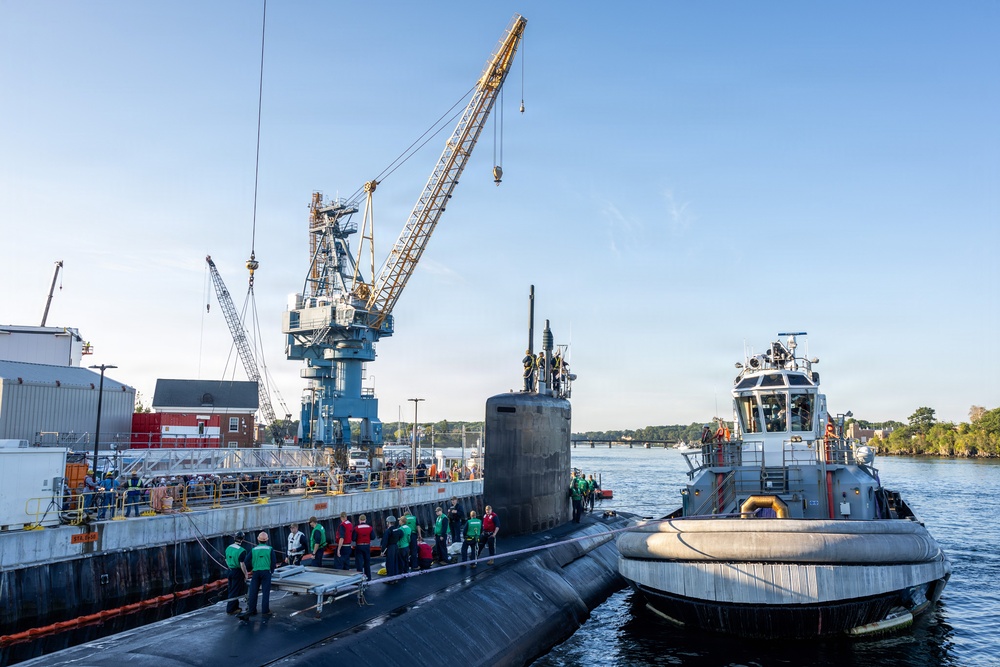 A submarine is being docked or maneuvered at a naval shipyard facility, assisted by a tugboat on its starboard side. Numerous personnel in green and red vests are gathered on the dock and submarine hull, suggesting a significant arrival or departure event. Large industrial cranes are visible in the background, indicating an active shipyard environment.