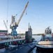 A submarine is being docked or maneuvered at a naval shipyard facility, assisted by a tugboat on its starboard side. Numerous personnel in green and red vests are gathered on the dock and submarine hull, suggesting a significant arrival or departure event. Large industrial cranes are visible in the background, indicating an active shipyard environment.