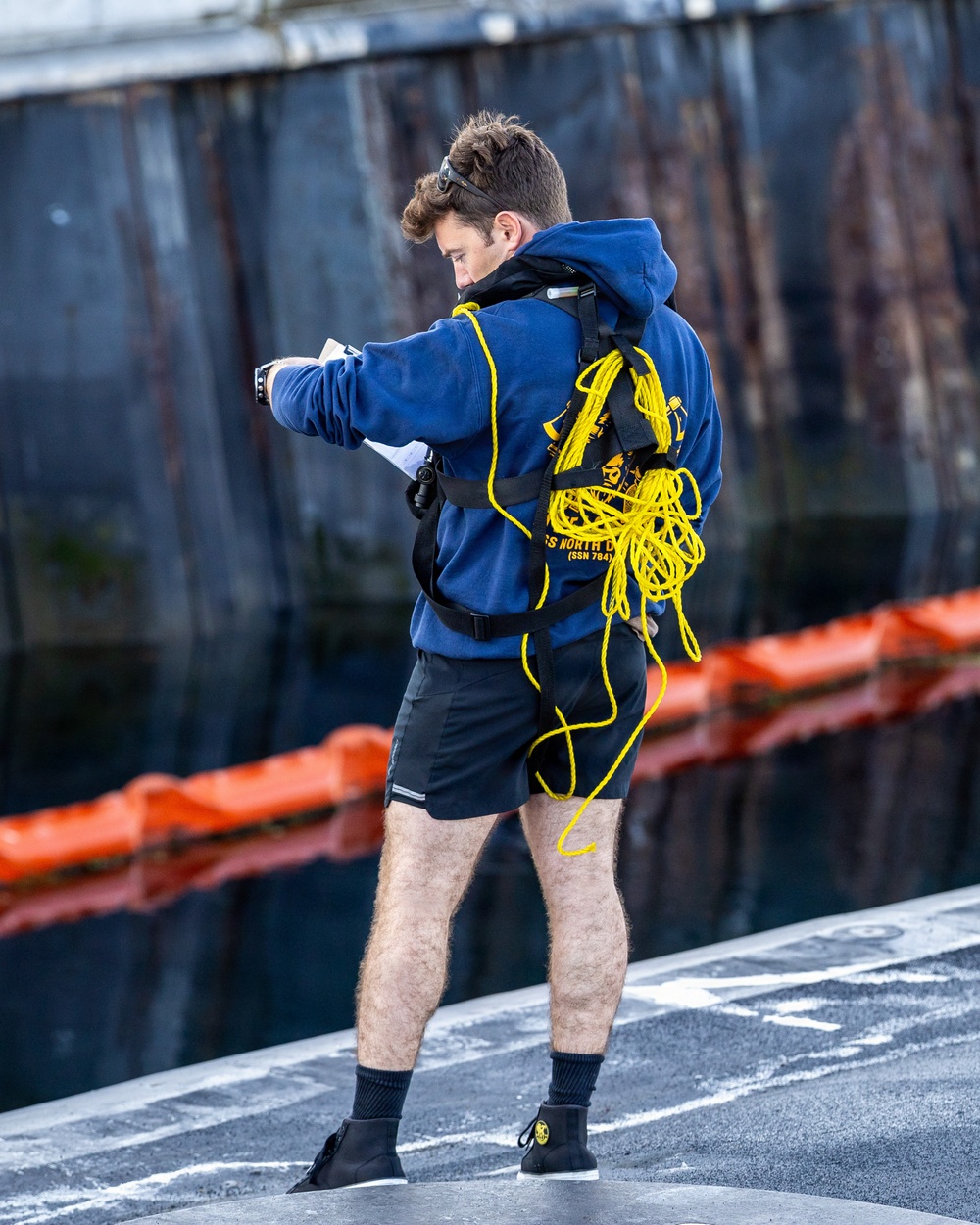 A young man stands on what appears to be the deck of a submarine or naval vessel, wearing a blue hoodie with 'USS NORTH DAKOTA (SSN 784)' printed on the back. He is equipped with a black safety harness carrying a coiled yellow rope, and appears to be looking at or operating a small device in his hands. Orange safety barriers and a dark metal hull are visible in the background.
