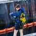 A young man stands on what appears to be the deck of a submarine or naval vessel, wearing a blue hoodie with 'USS NORTH DAKOTA (SSN 784)' printed on the back. He is equipped with a black safety harness carrying a coiled yellow rope, and appears to be looking at or operating a small device in his hands. Orange safety barriers and a dark metal hull are visible in the background.