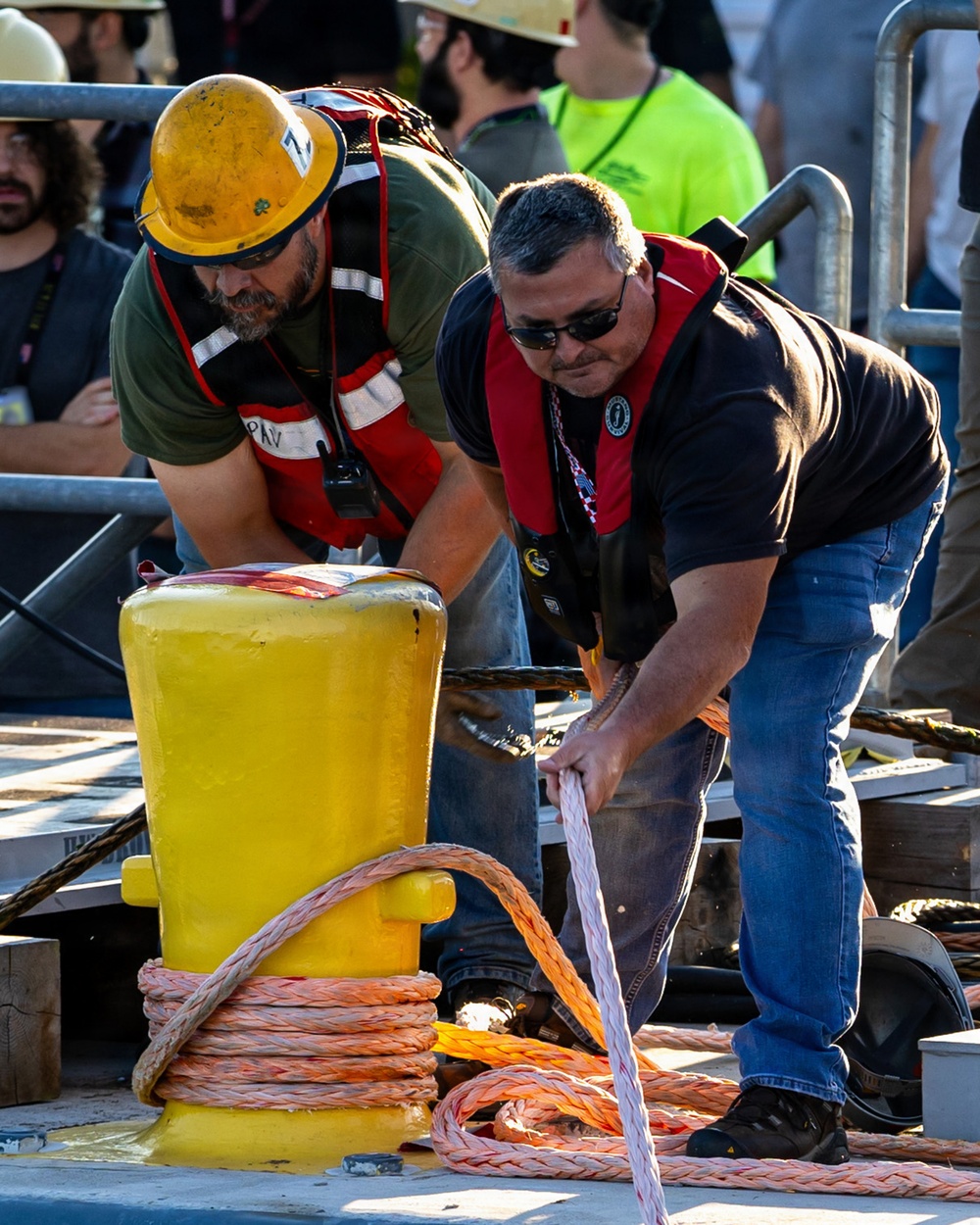 Two workers are handling mooring lines on what appears to be a dock or vessel deck, working together around a large yellow painted bollard. Both men are wearing red inflatable personal flotation devices (PFDs), indicating a maritime safety environment. A crowd of observers and additional workers in safety gear are visible in the background.