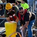 Two workers are handling mooring lines on what appears to be a dock or vessel deck, working together around a large yellow painted bollard. Both men are wearing red inflatable personal flotation devices (PFDs), indicating a maritime safety environment. A crowd of observers and additional workers in safety gear are visible in the background.