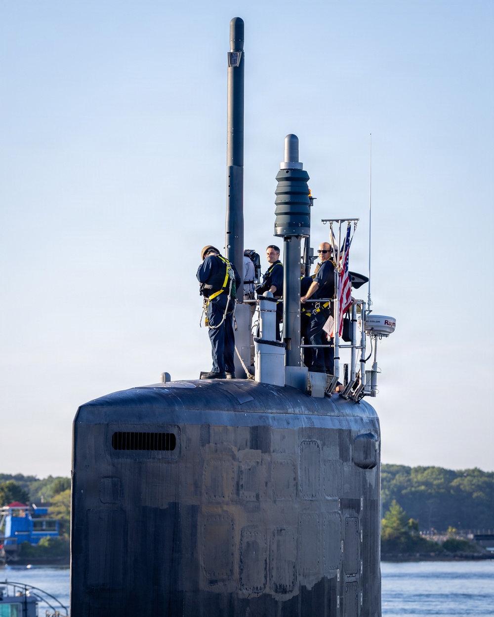 The image shows the sail (conning tower) of a U.S. Navy submarine with crew members stationed on top during what appears to be a port arrival or departure. Four personnel in dark navy uniforms with yellow safety harnesses are visible on the bridge and hull. An American flag is displayed on the sail, and various mast-mounted sensors and antennas are visible.