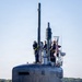 The image shows the sail (conning tower) of a U.S. Navy submarine with crew members stationed on top during what appears to be a port arrival or departure. Four personnel in dark navy uniforms with yellow safety harnesses are visible on the bridge and hull. An American flag is displayed on the sail, and various mast-mounted sensors and antennas are visible.