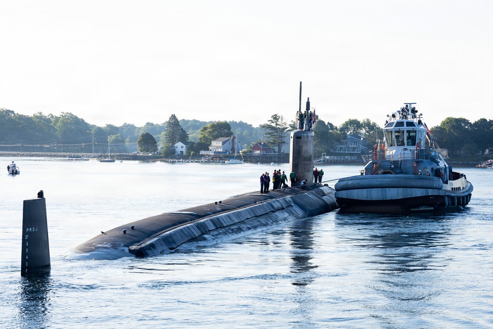A large military submarine is navigating a calm waterway, assisted by a tugboat positioned alongside its starboard side. Several crew members are visible standing on the submarine's hull near the sail (conning tower). The scene appears to be a harbor or river environment with residential buildings and dense tree lines visible in the background.