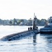 A large military submarine is navigating a calm waterway, assisted by a tugboat positioned alongside its starboard side. Several crew members are visible standing on the submarine's hull near the sail (conning tower). The scene appears to be a harbor or river environment with residential buildings and dense tree lines visible in the background.