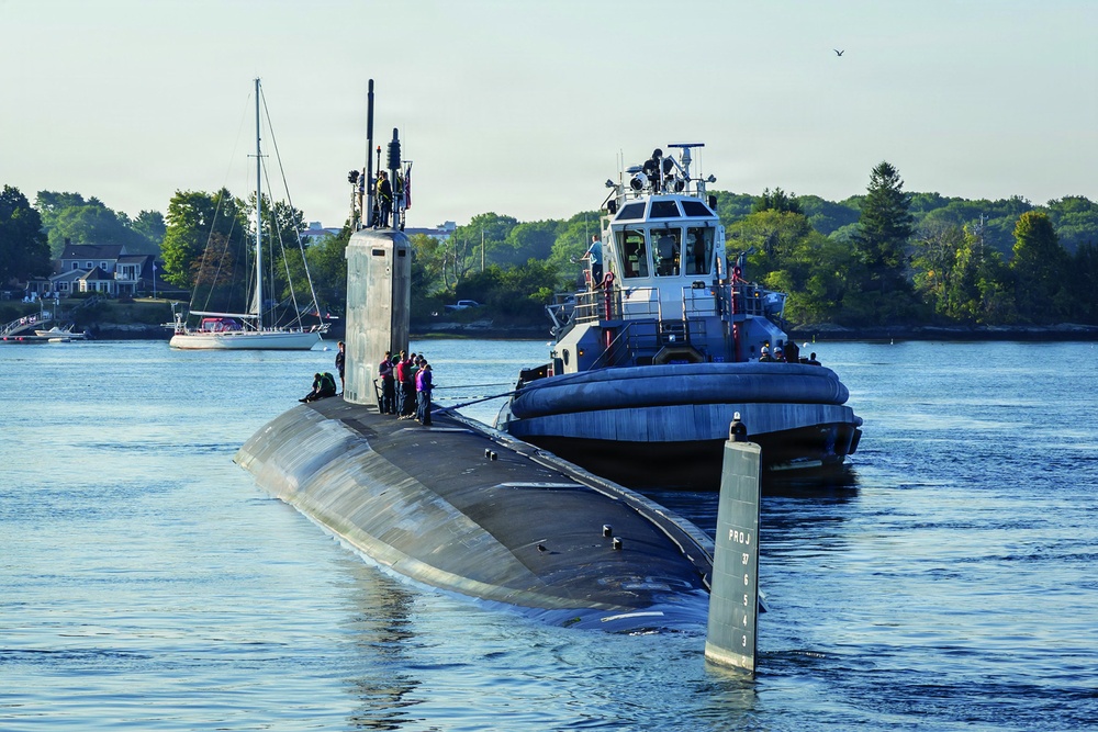 A military submarine is navigating a river or harbor channel accompanied by a tugboat positioned alongside its starboard side. Several personnel are visible on the submarine's deck and sail (conning tower), suggesting the vessel is either departing or arriving at port. The scene is set in a calm, tree-lined waterway with civilian sailboats visible in the background.