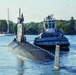 A military submarine is navigating a river or harbor channel accompanied by a tugboat positioned alongside its starboard side. Several personnel are visible on the submarine's deck and sail (conning tower), suggesting the vessel is either departing or arriving at port. The scene is set in a calm, tree-lined waterway with civilian sailboats visible in the background.