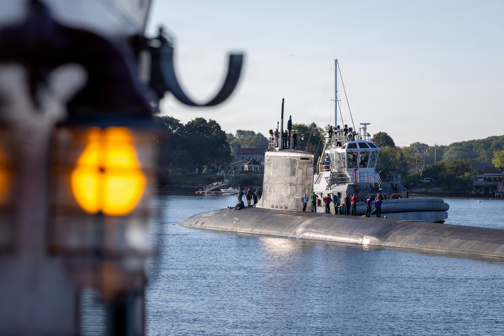 A submarine is navigating a calm waterway, accompanied by a tugboat positioned alongside its hull. Multiple personnel are visible on the submarine's deck and sail (conning tower), suggesting an arrival or departure operation. The scene is photographed from another vessel, with a blurred amber navigation light visible in the foreground.