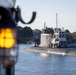 A submarine is navigating a calm waterway, accompanied by a tugboat positioned alongside its hull. Multiple personnel are visible on the submarine's deck and sail (conning tower), suggesting an arrival or departure operation. The scene is photographed from another vessel, with a blurred amber navigation light visible in the foreground.