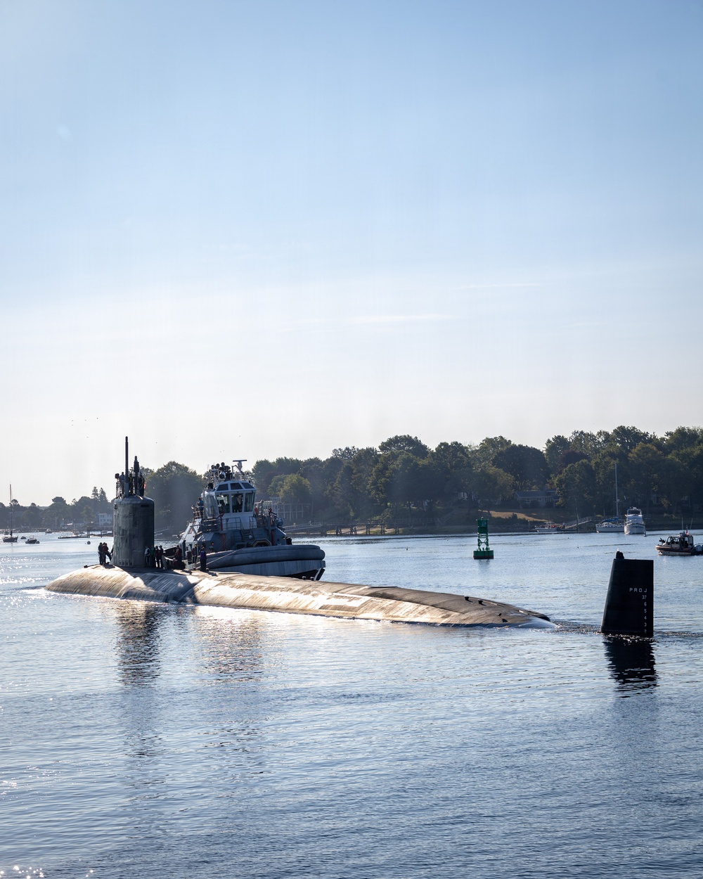 A military submarine is navigating a harbor channel assisted by a tugboat positioned alongside its hull. Personnel are visible on the submarine's deck and sail (conning tower). The scene takes place in calm, sunlit waters with a tree-lined shoreline and navigational buoys visible in the background.