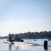 A military submarine is navigating a harbor channel assisted by a tugboat positioned alongside its hull. Personnel are visible on the submarine's deck and sail (conning tower). The scene takes place in calm, sunlit waters with a tree-lined shoreline and navigational buoys visible in the background.