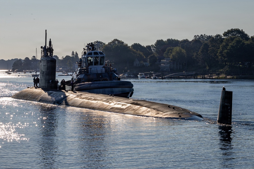 A military submarine is navigating a calm waterway, assisted by a tugboat positioned alongside its hull. Several crew members are visible on the submarine's deck near the sail (conning tower). The scene is set in what appears to be a coastal or river inlet, with tree-lined shores and residential structures visible in the background.
