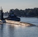 A military submarine is navigating a calm waterway, assisted by a tugboat positioned alongside its hull. Several crew members are visible on the submarine's deck near the sail (conning tower). The scene is set in what appears to be a coastal or river inlet, with tree-lined shores and residential structures visible in the background.
