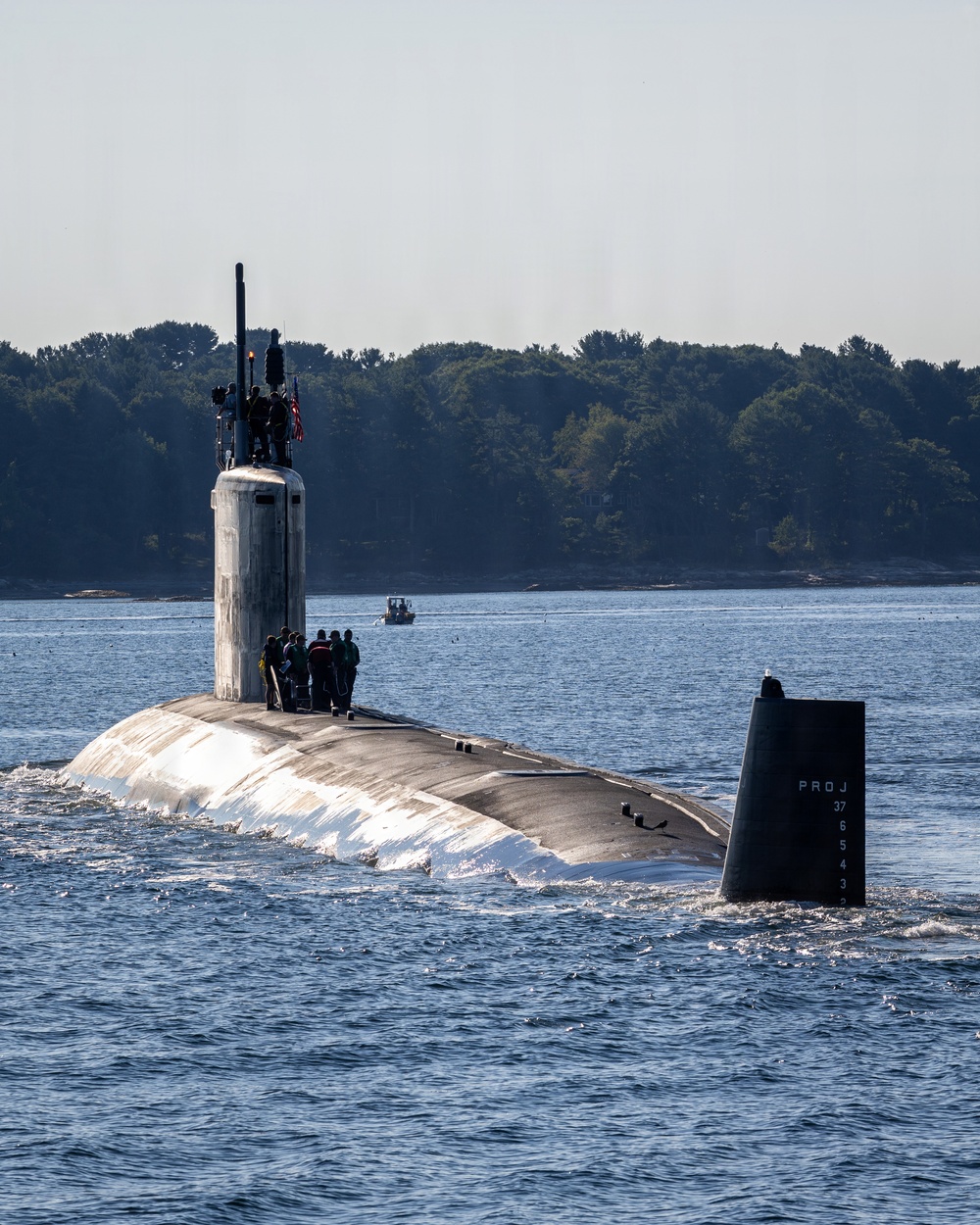 A U.S. Navy submarine is underway on the surface of a calm waterway, with crew members visible on the deck and atop the sail (conning tower). An American flag is visible on the sail, and depth markings labeled 'PROJ' with numbers (37, 6, 5, 4, 3) are stenciled on the stern rudder/fin. A small civilian or escort vessel is visible in the background near a tree-lined shoreline.