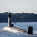 A U.S. Navy submarine is underway on the surface of a calm waterway, with crew members visible on the deck and atop the sail (conning tower). An American flag is visible on the sail, and depth markings labeled 'PROJ' with numbers (37, 6, 5, 4, 3) are stenciled on the stern rudder/fin. A small civilian or escort vessel is visible in the background near a tree-lined shoreline.