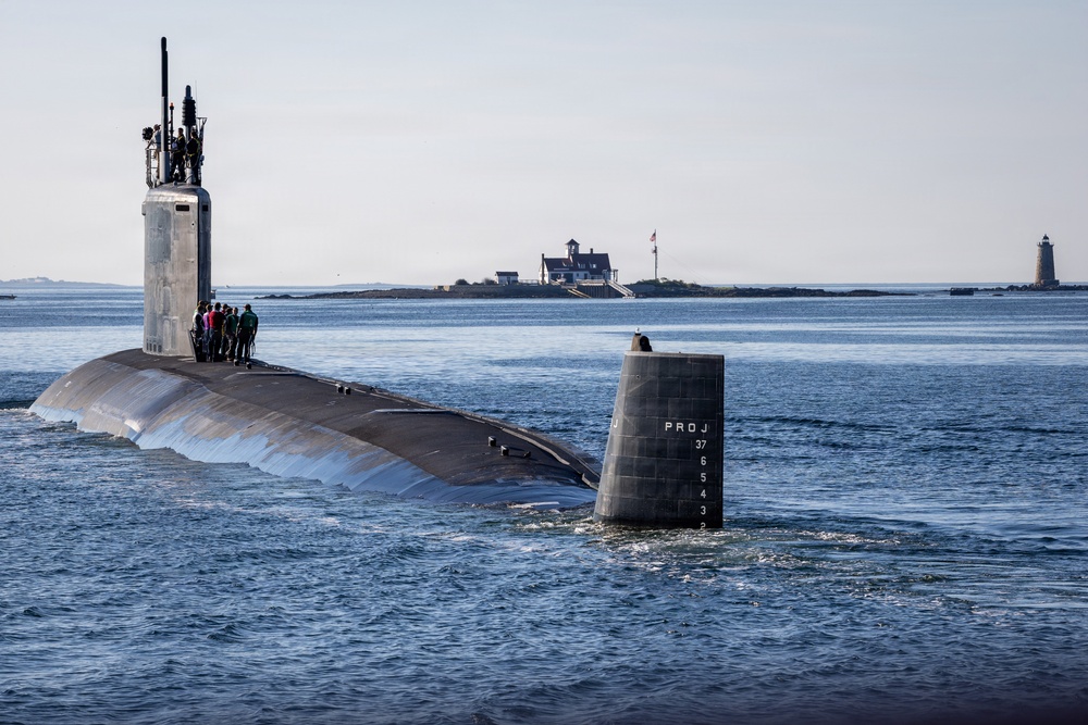 A large military submarine is navigating through open coastal waters with several crew members visible on the forward deck near the sail (conning tower). The submarine's stern section displays markings including 'PROJ' and numerical depth/draft indicators (2-7). In the background, a small island with buildings and a flagpole is visible, along with a lighthouse further in the distance.